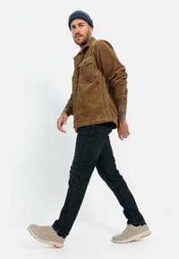 Een corduroy jas in bruin, donkere spijkerbroek en beige sneakers. De jas heeft twee borstzakken; de outfit is casual en functioneel.