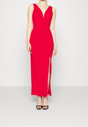 Femme portant une robe rouge sans manches, longue jusqu'au sol, avec un col en V et une fente latérale, associée à des talons ouverts noirs, debout devant un fond uni.