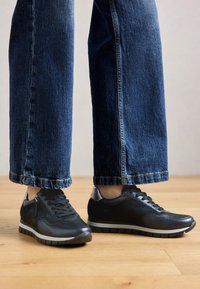 Baskets basses noires en cuir et tissu, avec une surface texturée, une semelle grise contrastante et un accent brillant au niveau du talon.