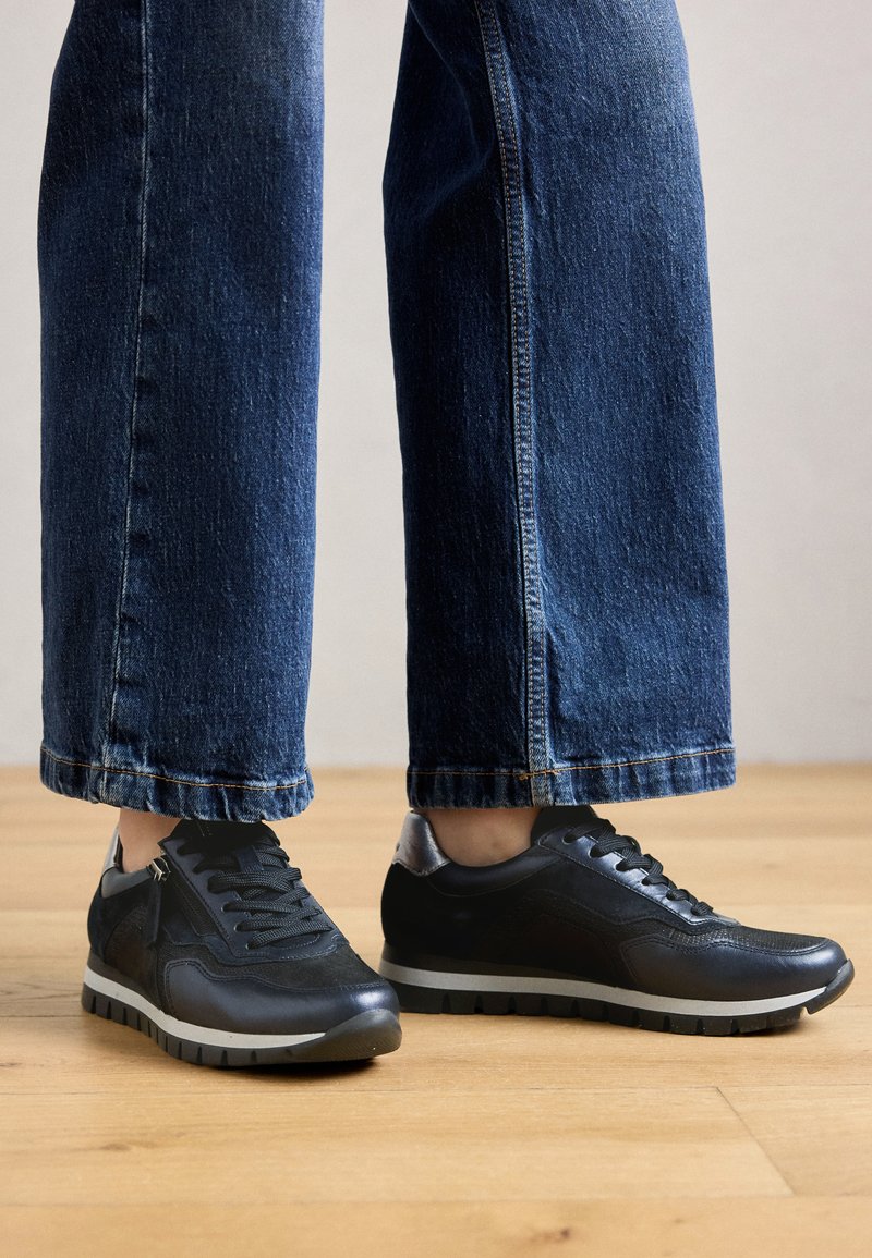 Baskets basses noires en cuir et tissu, avec une surface texturée, une semelle grise contrastante et un accent brillant au niveau du talon.