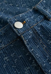 Povečava teksturirane modre denim tkanine z oranžnim šivom in srebrnim kovinskim gumbom na šivu oblačila.