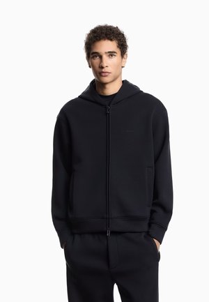 Felpa nera con zip realizzata in un tessuto morbido, dotata di tasca frontale, cappuccio regolabile e polsini a coste. Design minimalista senza logo visibili.