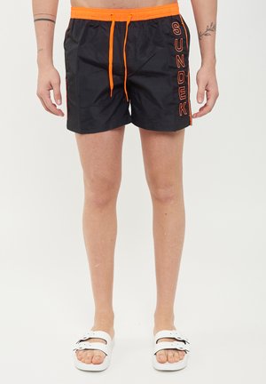 Pantaloni da bagno neri con coulisse arancione e dettaglio del logo "SUNDEK". Realizzati in tessuto morbido, offrono una vestibilità comoda e una lunghezza sopra il ginocchio.