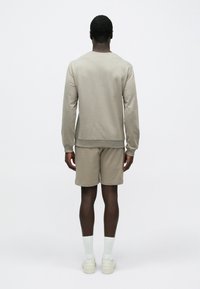 Egy férfi áll háttal nekünk, bézs hosszú ujjú felsőt és rövidnadrágot visel, fehér zoknival és fehér tornacipővel, egyszínű világos háttér előtt.