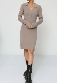 Robe tricotée en brun clair avec une texture côtelée, des manches longues, un décolleté en V et des accents de boutons. Assortie avec des bottines noires à la cheville.