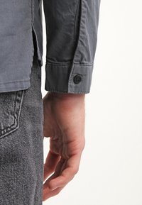 Camisa gris de manga larga con puños abotonados, que presenta una textura suave y un ligero brillo. Costuras visibles y un detalle de abertura lateral.