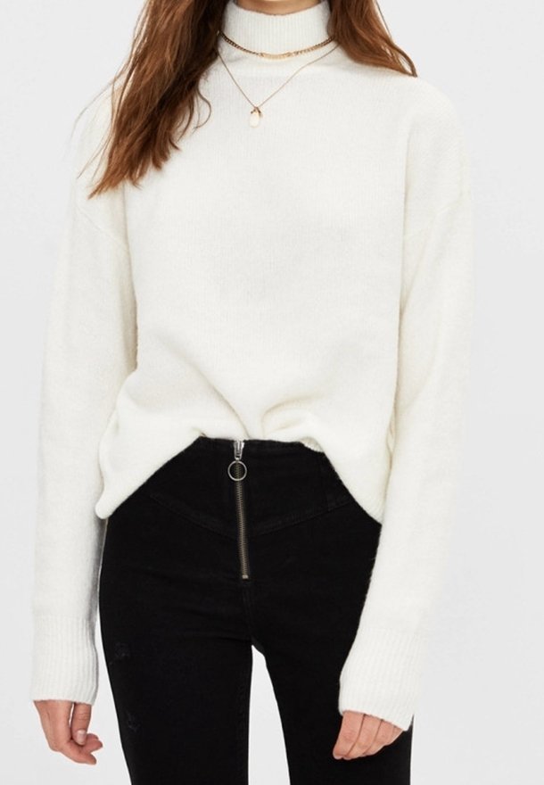Femme portant un pull à col roulé blanc, deux colliers dorés superposés et un pantalon noir taille haute avec une fermeture éclair devant et un anneau de tirage.