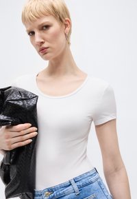 Ξανθιά γυναίκα με κοντά μαλλιά, που φοράει λευκό μπλουζάκι και μπλε τζιν, κρατάει ένα μαύρο υφή δερμάτινο τσαντάκι σε λευκό φόντο.