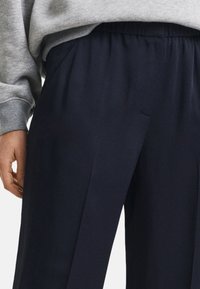 Pantaloni a gamba larga blu navy con cintura elastica, caratterizzati da una texture liscia e pieghe sottili sul davanti. Abbinati a una felpa grigia.