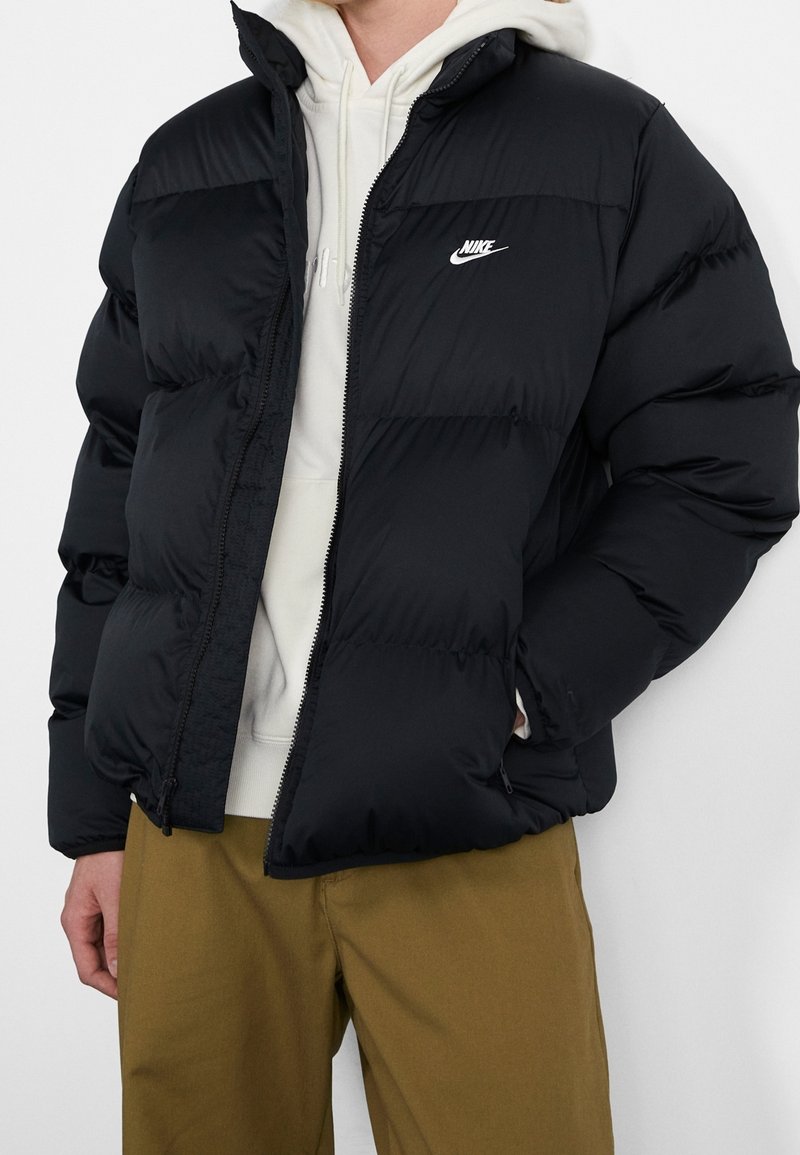 Persona vistiendo una chaqueta de plumas negra de Nike sobre una sudadera blanca con pantalones caqui, de pie contra un fondo blanco liso.