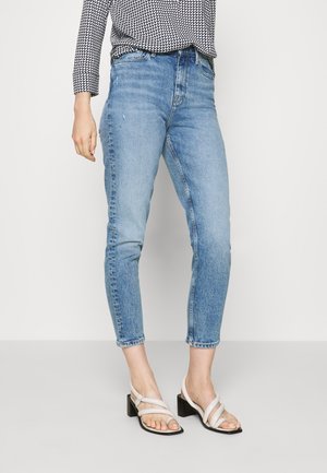 Kobieta ubrana w jasnoniebieskie obcisłe, wysokie spodnie jeansowe i białe sandały na obcasie, stojąca na jednolitym tle.