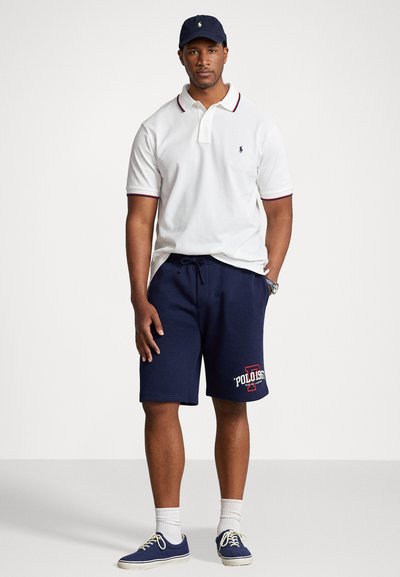 T-shirt polo blanche avec accents rouges et bleus marine, shorts de sudation bleus marine avec une impression rouge "Polo 1967", casquette bleue marine et baskets bleues.