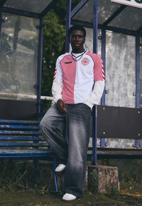 White and red striped jersey with logo, paired with gray wide-leg jeans and white sneakers. Model stands in a bus shelter setting.
