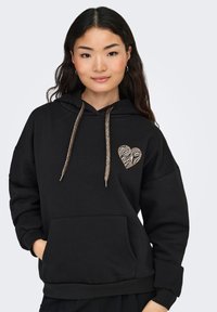 Sudadera negra con capucha que presenta un aplique en forma de corazón con patrón de cebra en el lado izquierdo del pecho, con cordones metálicos dorados y un bolsillo frontal.