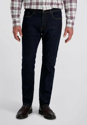 Mann in dunkelblauen Jeans, braunen Lederloafers und einem weißen Hemd mit rot-grauen Karomuster, der vor einem schlichten Hintergrund steht.