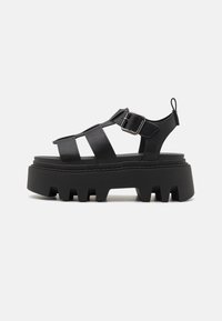 Black platform sandals with thick, ridged soles, featuring multiple strap designs and a side buckle closure for adjustable fit.