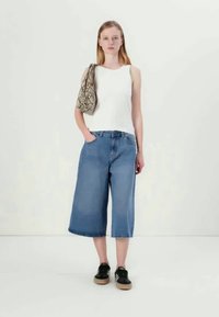 Λευκό αμάνικο μπλουζάκι, μπλε τζιν καμπάνα με ψηλή μέση και μαύρα αθλητικά παπούτσια. Σακίδιο ώμου με σχέδια. Το μοντέλο στέκεται μπροστά σε απλό φόντο.