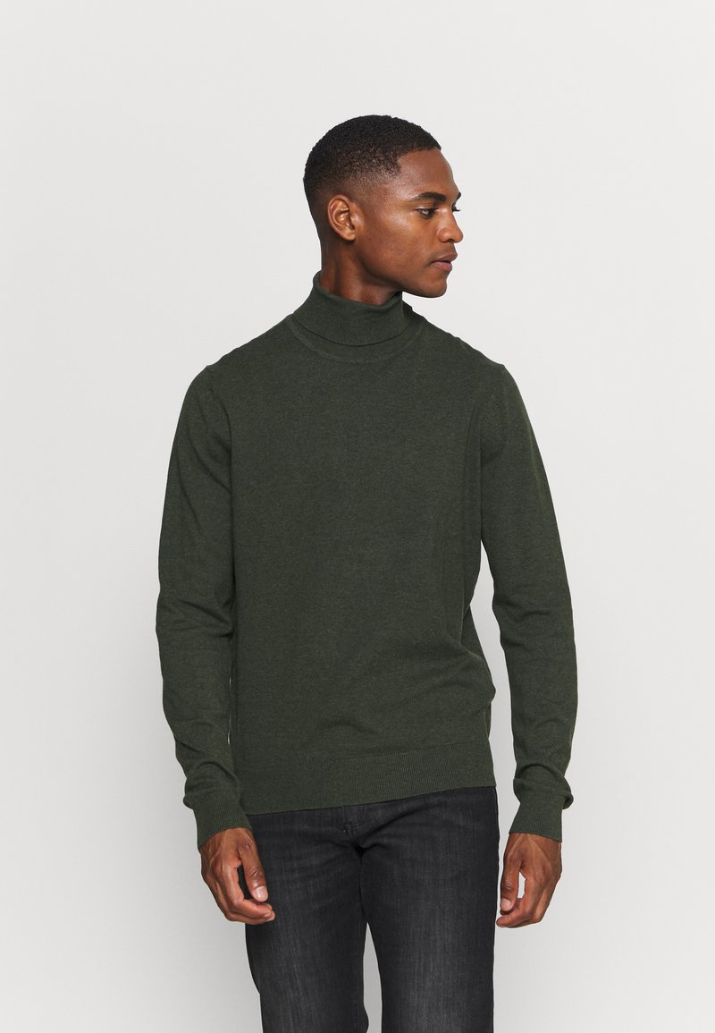 Dunkelgrüner Rollkragenpullover aus weichem Material mit enganliegenden Ärmeln und geripptem Saum, kombiniert mit dunklen Jeans. Seitenansicht des Models.