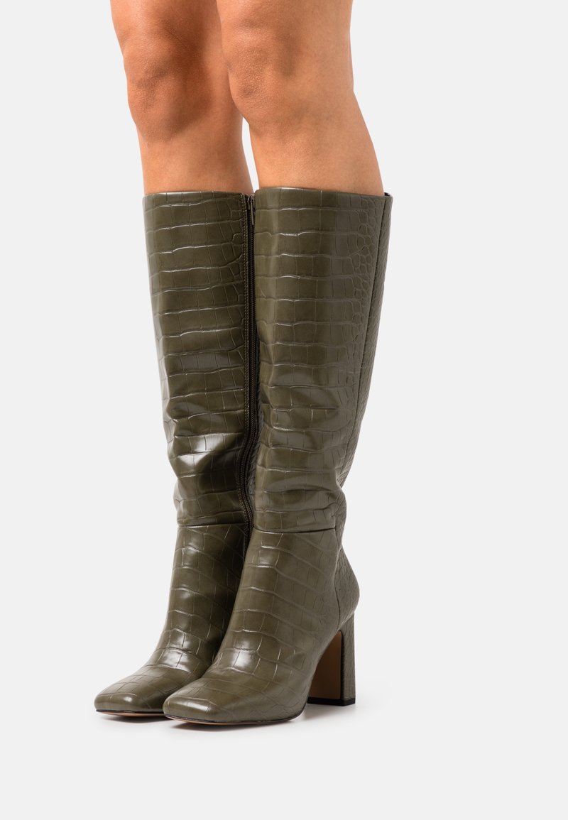 Jambes portant des bottes en cuir vert olive à motif crocodile, montantes jusqu'au genou, avec talons carrés et zips latéraux, sur fond blanc.