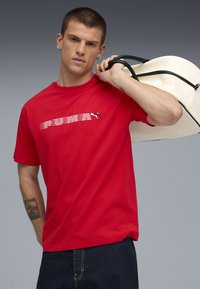 Rød bomulds T-shirt med hvid vandret "PUMA" logo, korte ærmer og en afslappet pasform. Model som bærer en hvid taske med sorte detaljer.