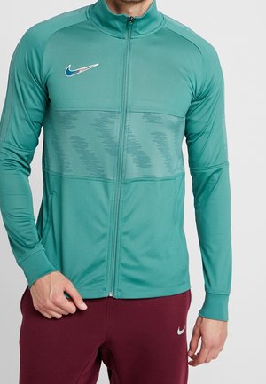 Hombre con chaqueta deportiva con cremallera en color verde azulado con logo azul de Nike y pantalones burdeos con el símbolo blanco de Nike, de pie frente a un fondo liso.