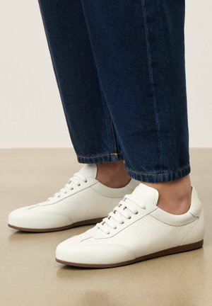 Biele nízke tenisky s textúrovanými bočnými panelmi nosené s tmavomodrými rifle s manžetou na hladkej béžovej podlahe.