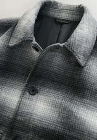 Grå ternet jakke med uldblandingsmateriale, krave og sort knaplukning. Indvendigt er den foret med en ensfarvet farve.