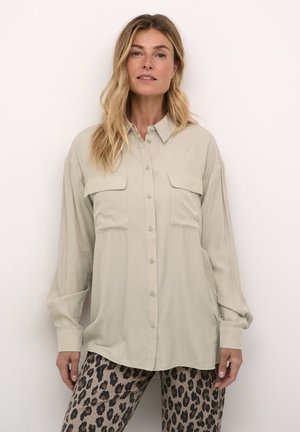 Vaalean beige nappipaita, jossa on kaksi rintataskua, löysä istuvuus ja pitkät hihat. Sileä kangas; kaareva helma laskeutuu kuvioitujen leopardiprinttipöksyjen päälle.