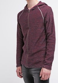 Röd och grå zip-up hoodie med en texturerad tyg, som har en huva och kontrasterande sömmar längs raglanärmarna.