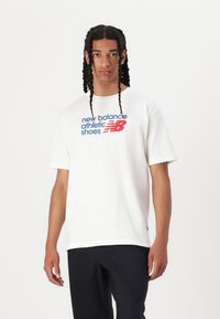 Fehér pamut póló, amelynek elütő kék és piros szövege van: "new balance athletic shoes" az elején, laza szabással és rövid ujjal.