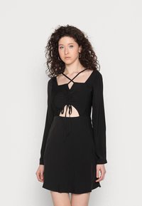 Vestido preto de mangas longas com decote quadrado, detalhes franzidos, uma seção central recortada com laços e saia ajustada. Textura de tecido suave.