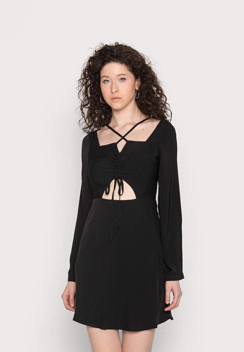 Vestido preto de mangas longas com decote quadrado, detalhes franzidos, uma seção central recortada com laços e saia ajustada. Textura de tecido suave.