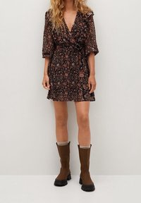 Robe portefeuille noire à fleurs avec de courtes manches bouffantes, taille cintrée et encolure à volants, associée à des bottes marron jusqu'aux genoux et à des socquettes grises.