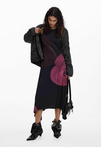 Czarna pikowana kurtka na ciemnej sukience z dużym fioletowym kwiatowym wzorem, zestawiona z czarnymi luźnymi botkami i małą torebką na ramię.