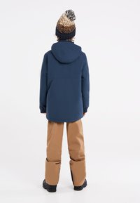 Veste imperméable marine avec capuche, bonnet en tricot à motifs crème et marine, et pantalon de ski beige, vu de derrière. Chaussures noires visibles.