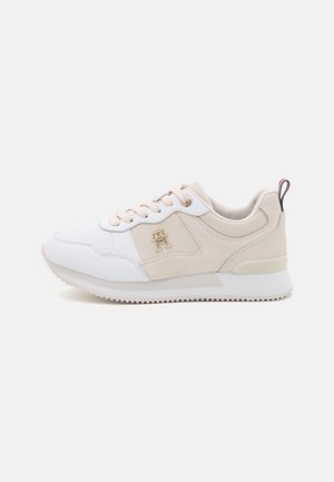 Baskets beige et blanches avec lacets, logo métallique de marque sur le côté, col rembourré et semelle en caoutchouc avec bande de roulement antidérapante.