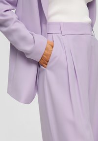 Lilac kostuum met een getailleerde blazer met een soepele textuur en iets te grote mouwen, gecombineerd met geplooide broeken.