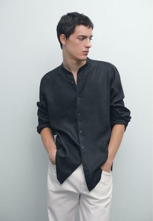 Camicia blu scuro con colletto alla cinese; maniche lunghe arrotolate sui polsini. Abbinata a pantaloni di colore chiaro. Tessuto liscio.