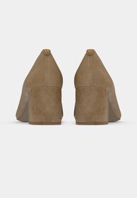 Beige ruskind sko med blokhæl, afrundet tå og gyldent logo på bagsiden. Glat tekstur og en mellemhøj hældesign.