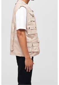 Beige arbetesväst i bomull med flera framfickor, dragkedja och tryckknappar. Bärs över en vit t-shirt, sidovy.