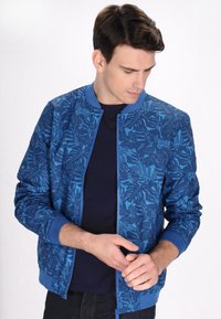 Giubbotto bomber blu con motivo a foglie, polsini a coste e chiusura con zip. Indossato sopra una maglietta navy, caratterizzata da una superficie liscia e un design leggero.