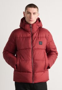 Crvena puffer jakna s kapuljačom, s horizontalno krojenim sekcijama, prednjim patentnim zatvaračem i crnim logom na lijevoj strani. Glatka tekstura.
