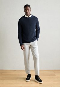 Marineblå ribbestrikket genser, grå skreddersydde bukser og svarte sneakers. Uformell antrekk med teksturert stoff og moderne passform. Nøytral bakgrunn.