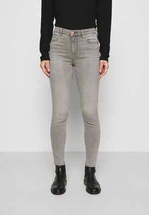 Osoba w jasnoszarych, wysokich skinny jeansach, czarnej bluzce z długim rękawem i czarnych botkach do kostek stoi na jednolitym tle.