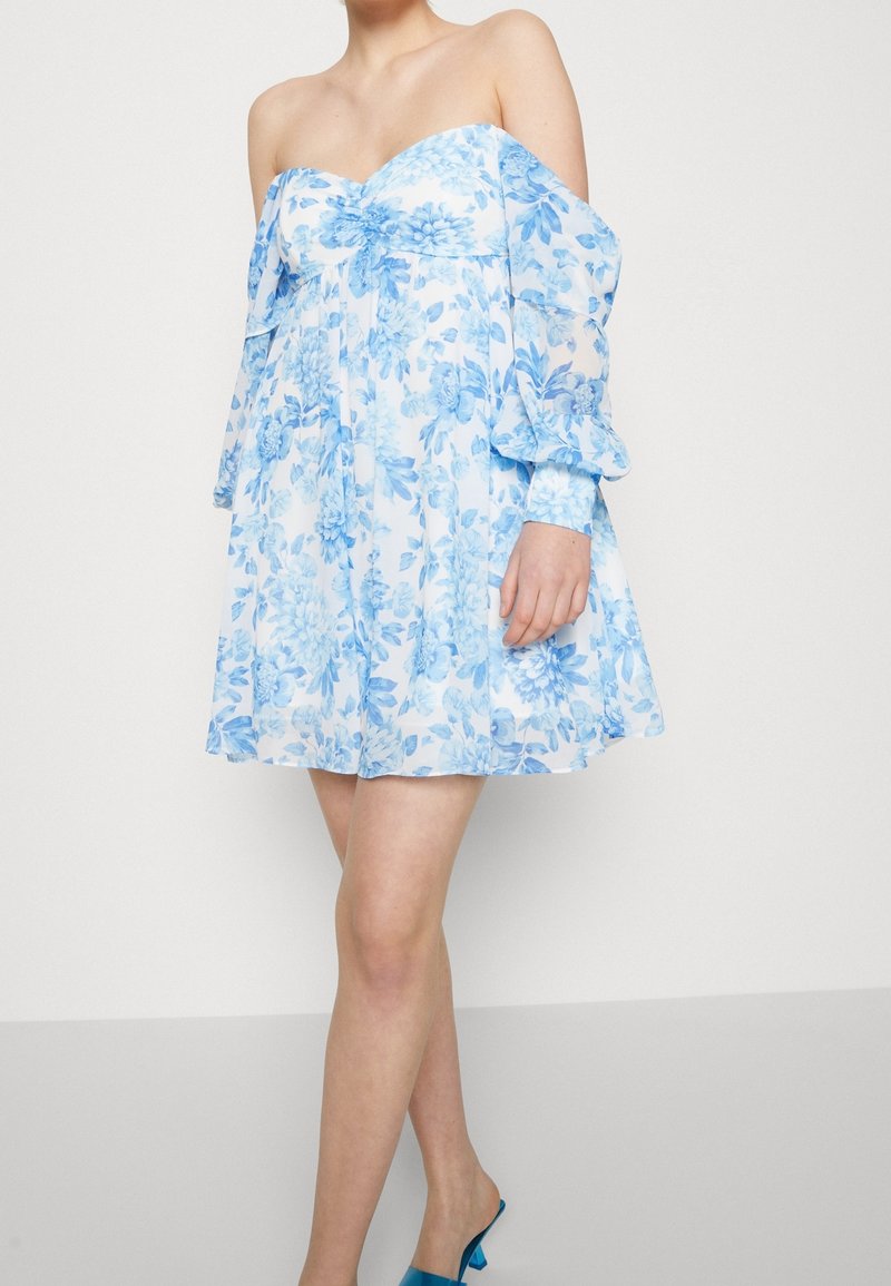 Femme portant une robe blanche à épaules dénudées et manches longues avec un imprimé floral bleu et des chaussures à talons bleus, debout devant un fond uni.