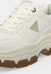 Baskets blanches Guess avec semelle extérieure épaisse marron avec rainures, panneau latéral matelassé, lacets et logo métallique triangulaire sur le côté.