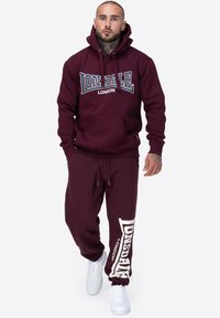 Burgundowa bluza z kapturem i pasujące dresy z nadrukiem "Lonsdale London" w białym kolorze. Wykonane z bawełny, z kieszenią typu kangur i elastycznym pasem. Białe trampki.