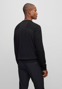 Črna pletena puloverja z rebrasto teksturo na ramenih in rokavih, z okroglo ovratnico in slim fit krojem. Obučen z črnimi hlačami.