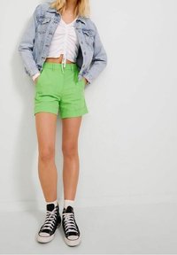 Pantaloni corti verdi con una vestibilità su misura, dotati di tasche laterali. Abbinati a un top corto bianco arricciato e una giacca di jeans, completati con sneakers nere.