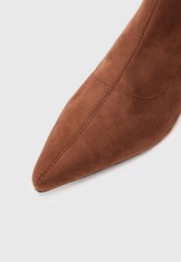 Scarpa in suede marrone a punta con una superficie liscia, caratterizzata da cuciture visibili lungo la parte superiore per un tocco di dettaglio. La suola è piatta con un accento minimale.
